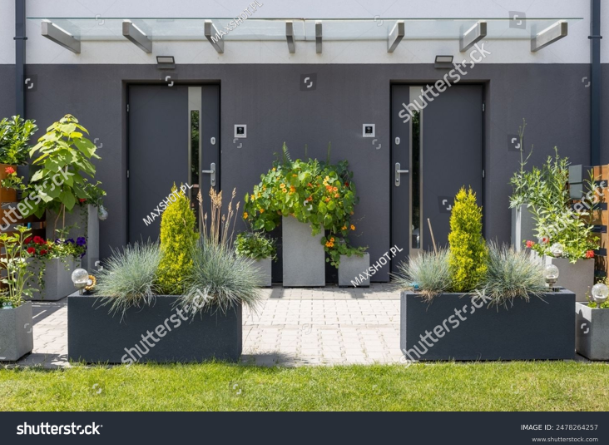 Modern apartment entrance with dark gray doors, decorative planters, colorful flowers, ornamental grasses, and a landscaped green lawn.